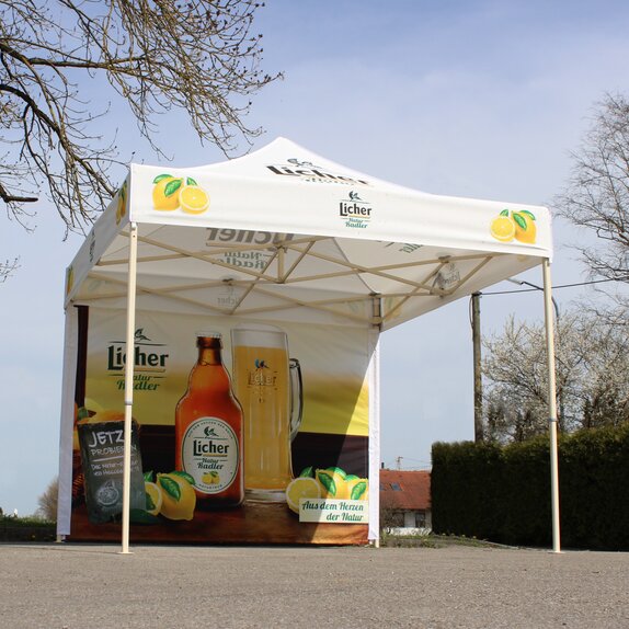 Ein voll-flächig bedruckter Faltpavillon, mit Fotos und dem Logo der Firma Licher Natur Radler, steht auf einer Straße neben einem Baum.