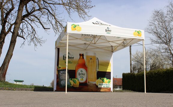 Ein voll-flächig bedruckter Faltpavillon, mit Fotos und dem Logo der Firma Licher Natur Radler, steht auf einer Straße neben einem Baum.