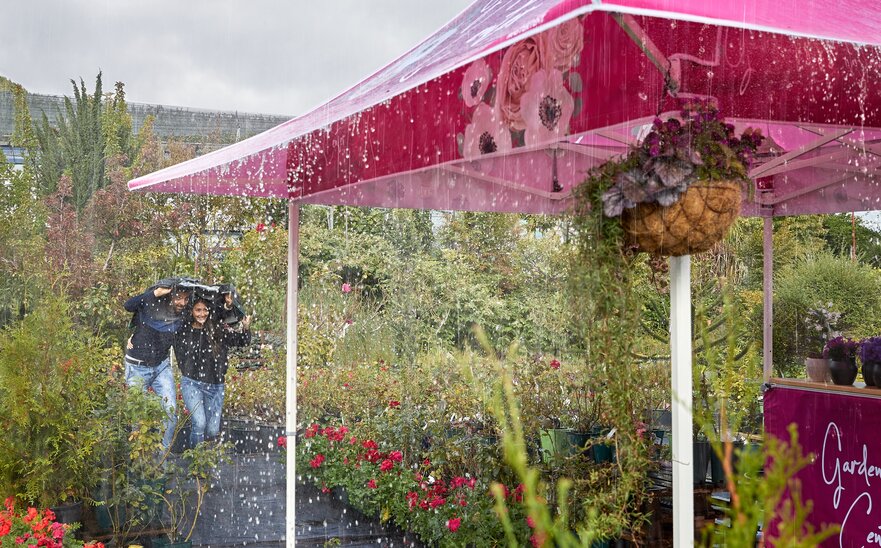 2 Personen, die im Regen mit einer Jacke über dem Kopf zum schutzbringenden Faltpavillon laufen