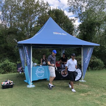 Two golfers are taking drinks at a blue gazebo with awnings and corner flags. 