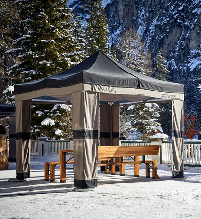 On a terrace there is a folding gazebo, underneath it a folding furniture. There is snow on the ground and on the trees and rocks in the background.