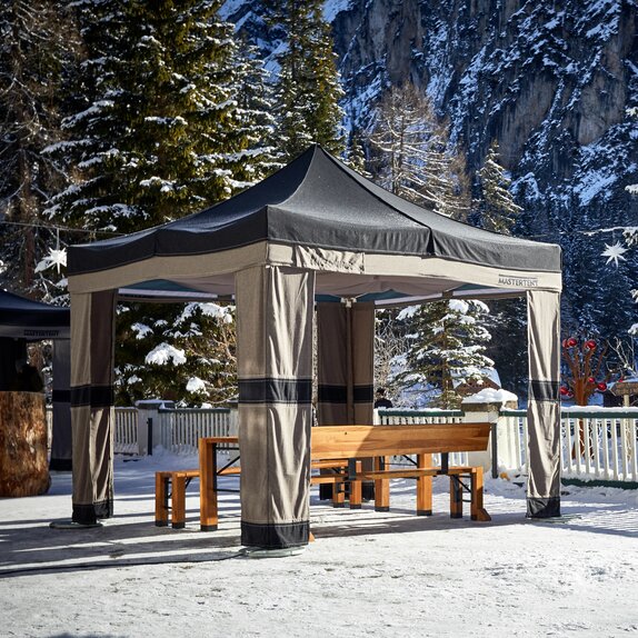 Auf einer Terrasse steht ein Faltpavillon, darunter eine Klappgarnitur. Am Boden liegt Schnee und im Hintergrund sieht man schneebedeckte Bäume und Felsen.