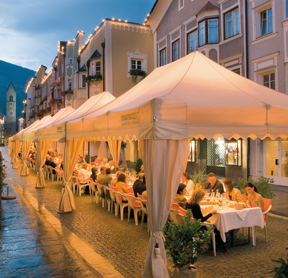 Set di gazebo esterni ecru eleganti 4x8m kit Royal con tende drappeggiate e frontalino ondulato per cena di lusso nella città di Vipiteno di sera con le luci Mastertent