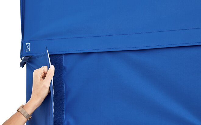 A woman is attaching the blue sidewall to the blue gazebo with a full-lenght velcro strip. 